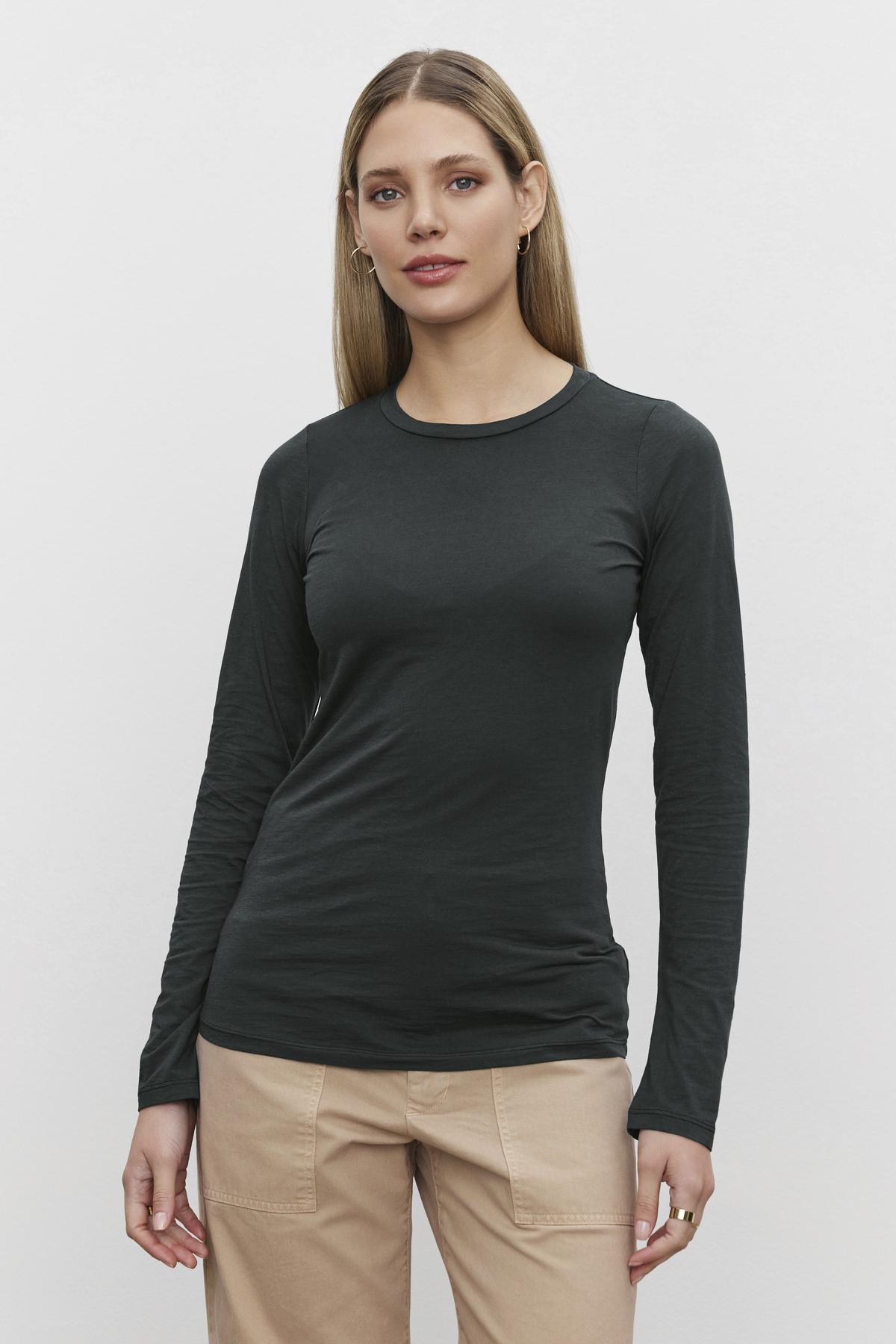   A person with long hair is wearing the ZOFINA TEE, a dark green fitted long sleeve top with a classic crew neckline from Velvet by Graham & Spencer, paired with beige pants while standing against a plain white background. 