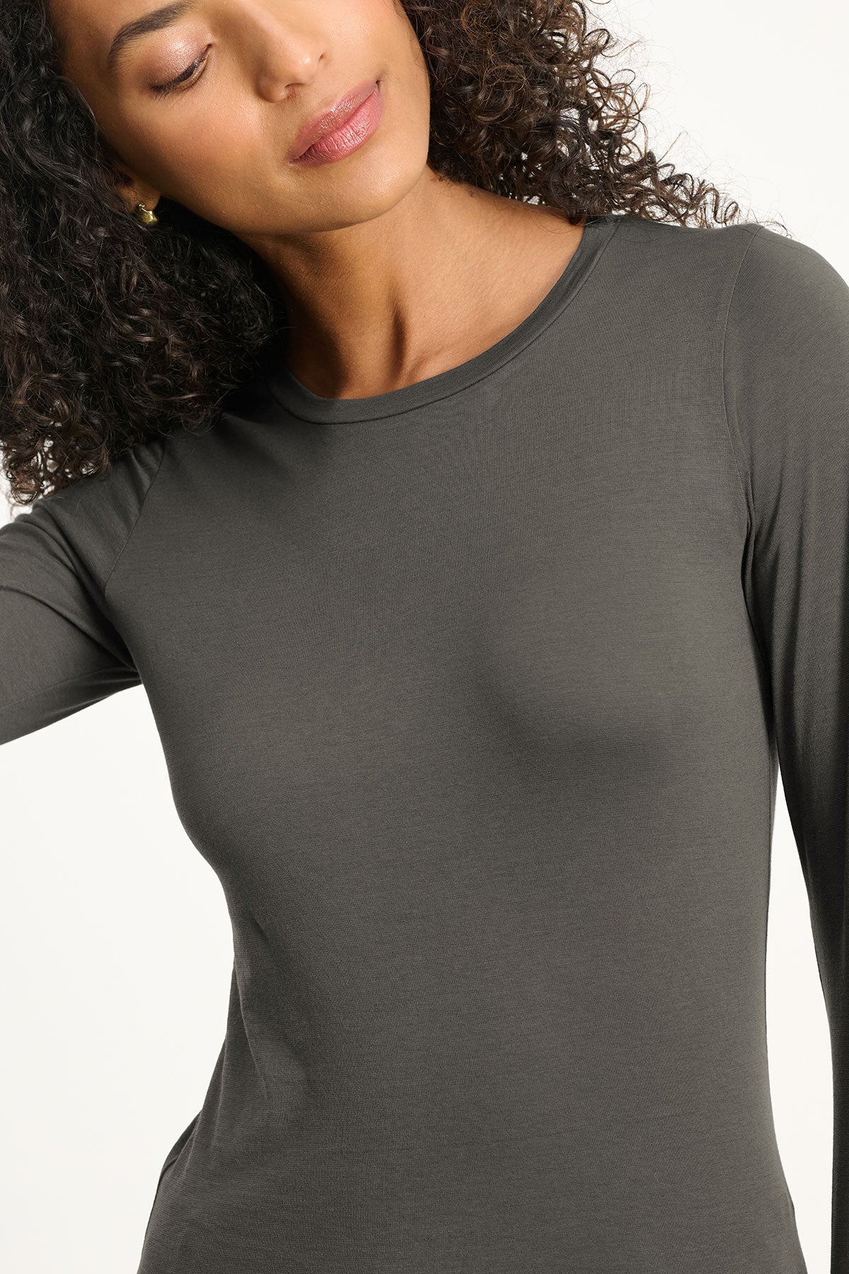 Woman with curly hair wears the ZOFINA TEE by Velvet by Graham & Spencer—an ultra-soft, fitted long sleeve from our basics collection—looking slightly downward against a plain background.-40417102495937