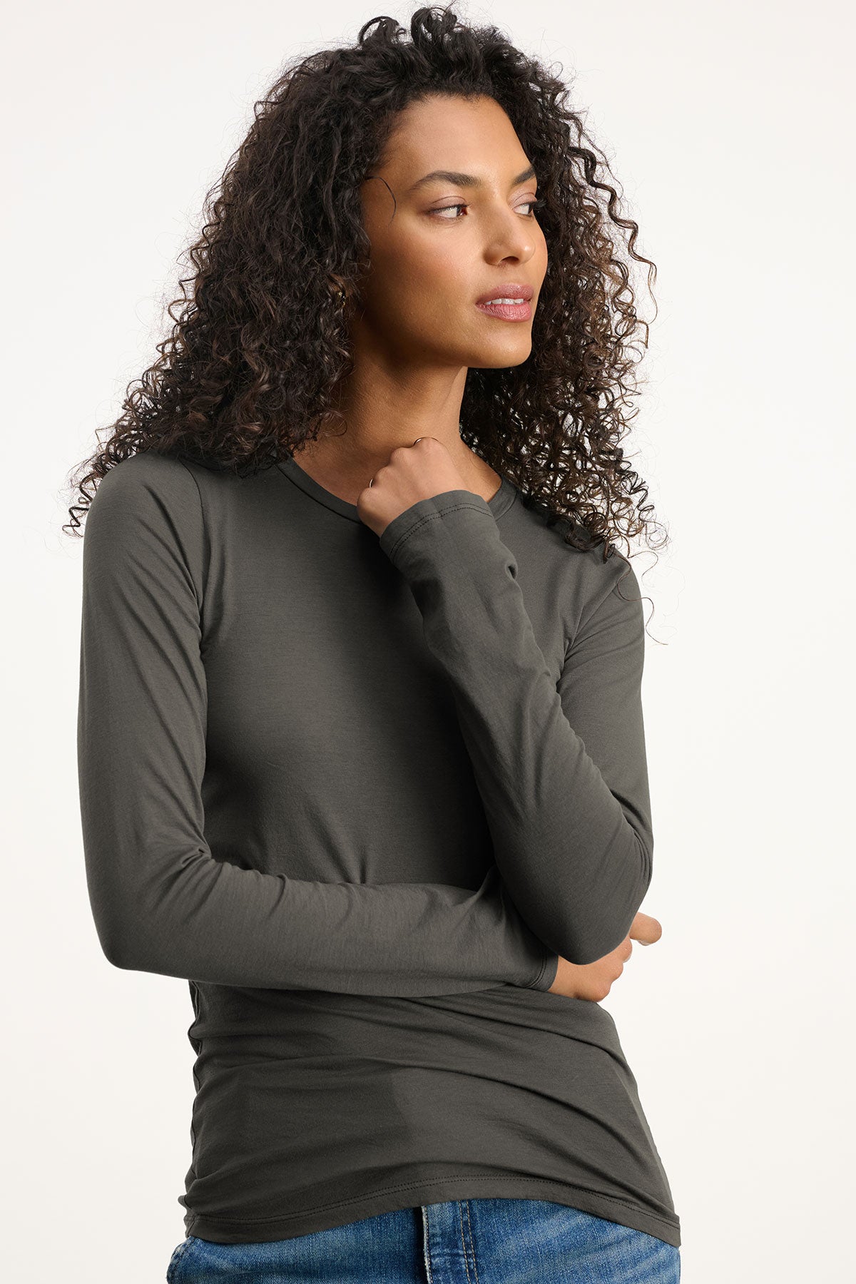 A woman with curly hair models the ZOFINA TEE by Velvet by Graham & Spencer—an ultra-soft, fitted long sleeve crew neck—paired with blue jeans against a plain light background, looking off to the side.-40417102364865