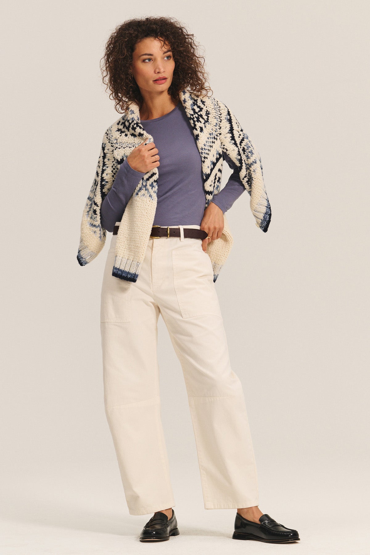 A woman stands in a relaxed pose wearing the ZOFINA TEE by Velvet by Graham & Spencer—a fitted, ultra-soft long sleeve with a classic crew neckline—paired with cream wide-leg pants, a brown belt, black loafers, and a patterned knit shawl.