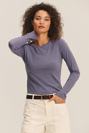 A woman with curly hair wears the ZOFINA TEE by Velvet by Graham & Spencer—an ultra-soft, fitted long sleeve in purple—paired with white pants and a brown belt, posing with one hand in her pocket against a plain background.