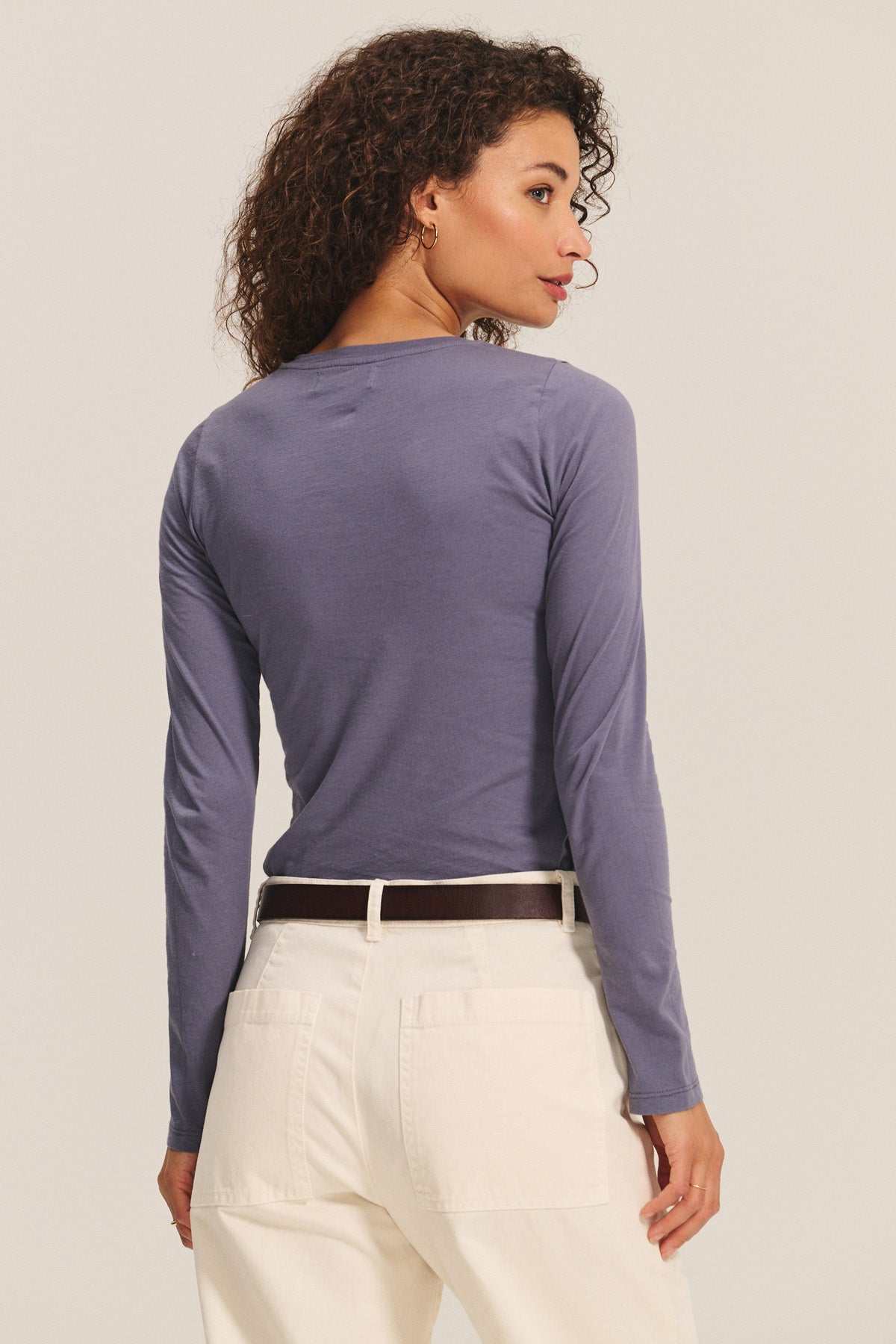A woman with curly hair wears the ZOFINA TEE by Velvet by Graham & Spencer, a fitted long sleeve crew neck, paired with white pants and stands with her back turned, glancing over her shoulder.-40732698804417