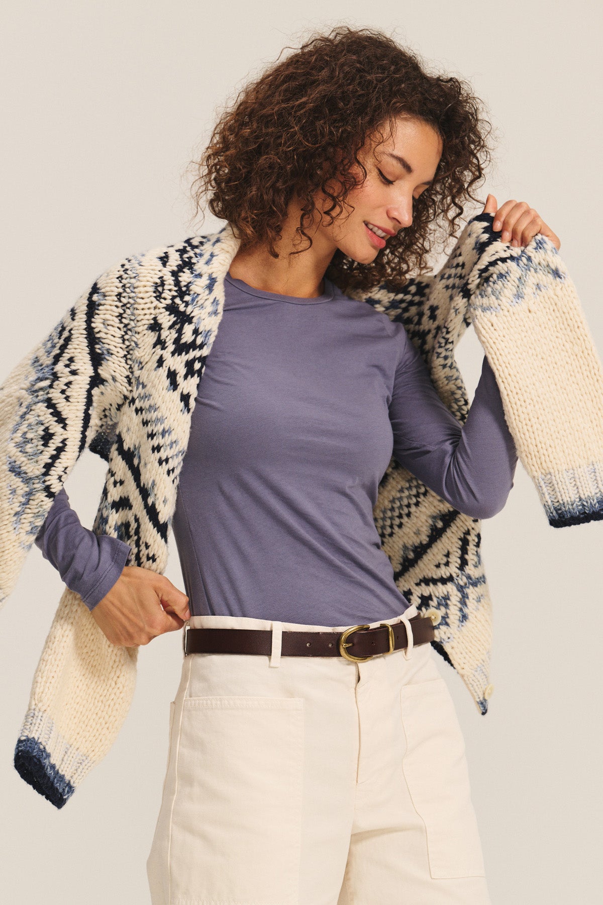 A woman with curly hair wears the Velvet by Graham & Spencer ZOFINA TEE, a blue fitted long sleeve crewneck, styled with cream pants and a patterned knit cardigan draped over her shoulders as she looks down.-40732698738881