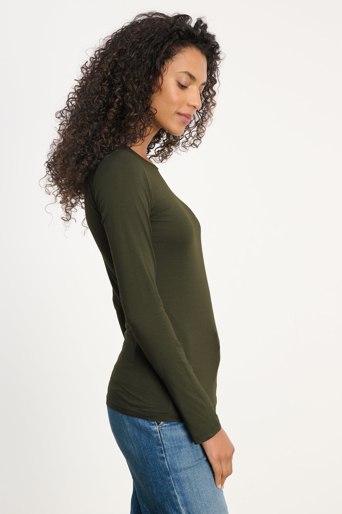 A person with long curly hair wears the ZOFINA TEE by Velvet by Graham & Spencer—an ultra-soft, fitted long sleeve with a classic crew neckline—paired with blue jeans, standing in profile against a plain light background.-40182503801025