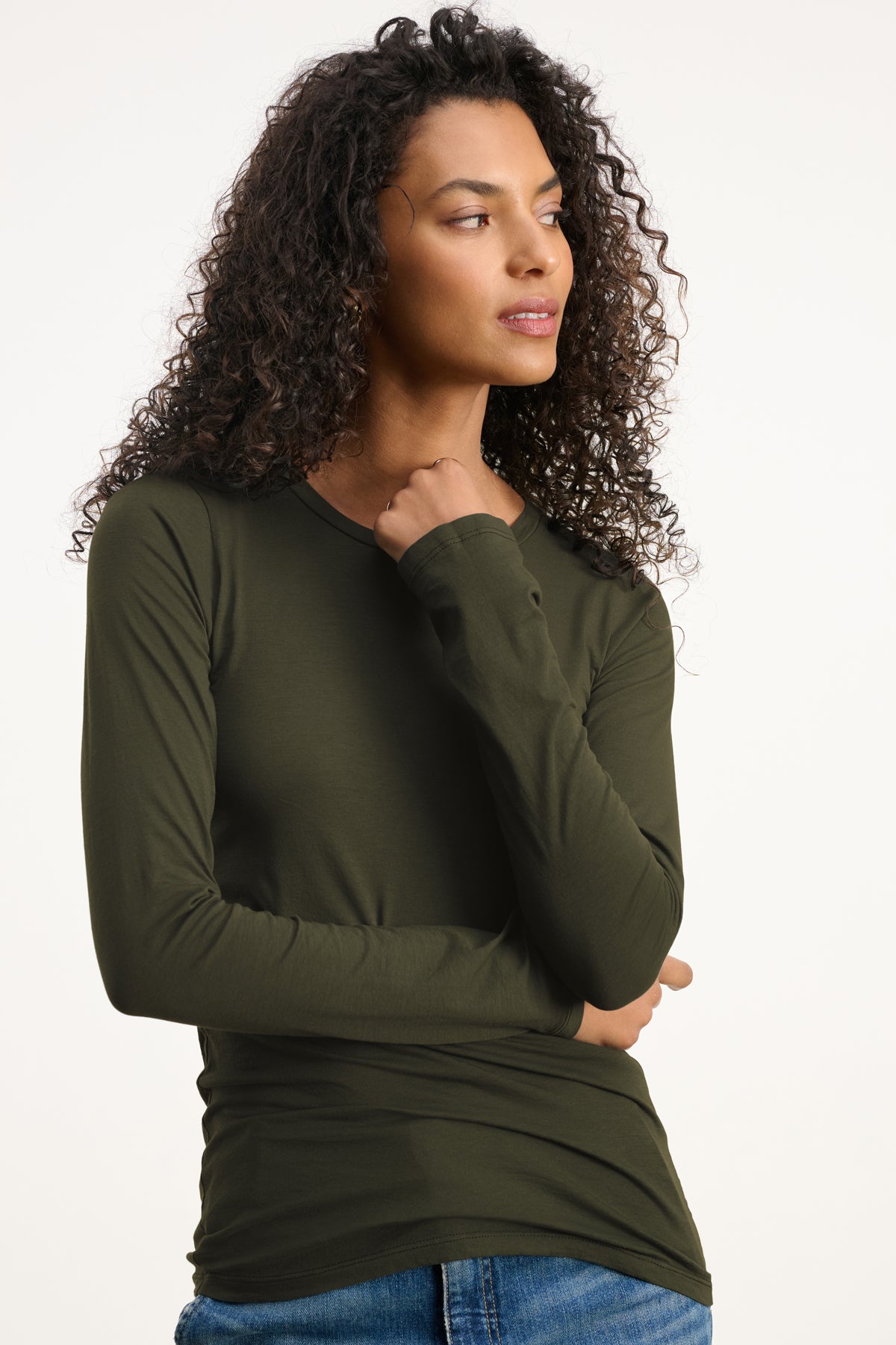   A woman with curly hair wears the ZOFINA TEE by Velvet by Graham & Spencer in olive green with blue jeans, standing against a plain background and looking to her right. 