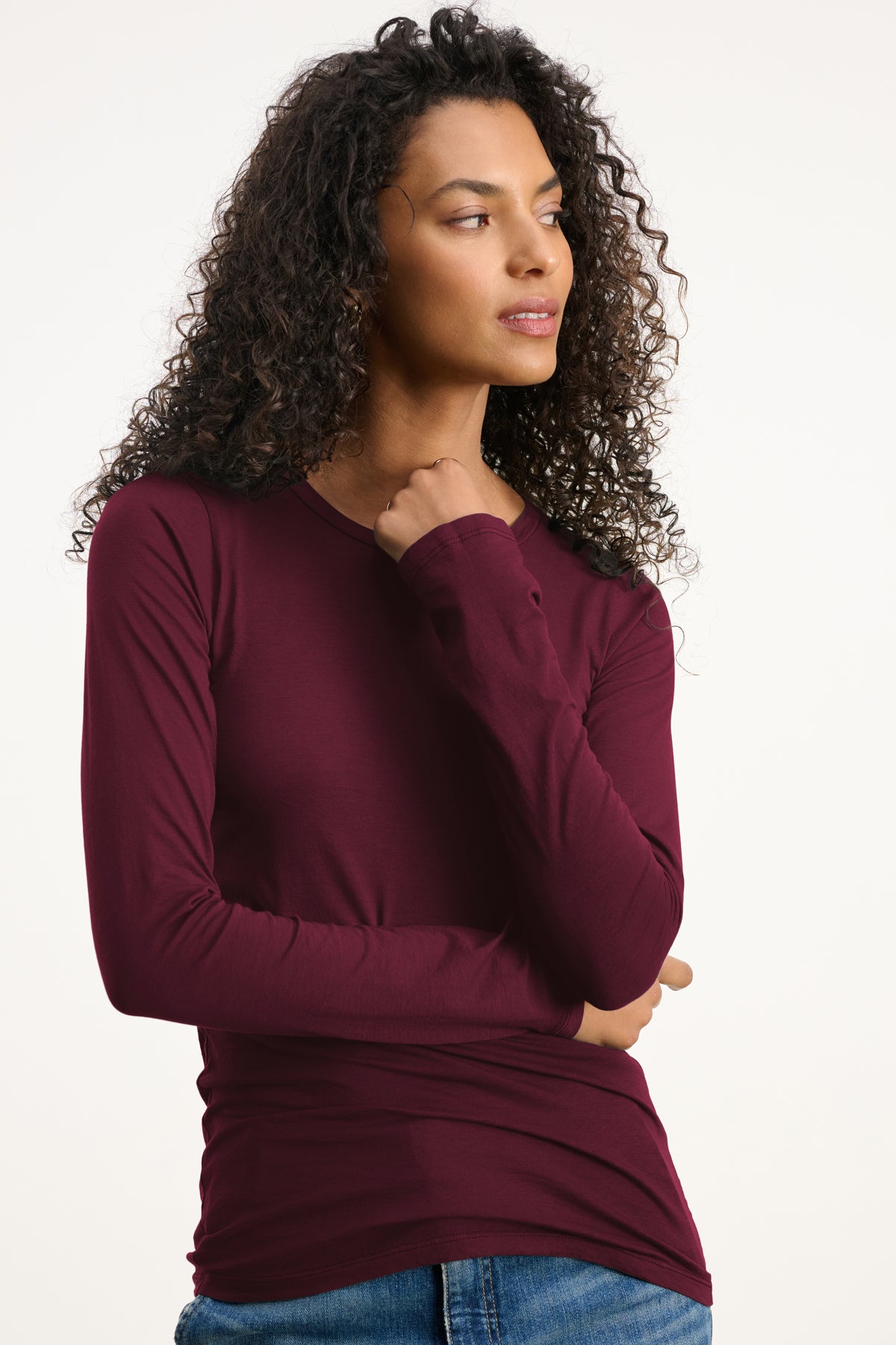 A person with long curly hair wears the ZOFINA TEE by Velvet by Graham & Spencer—a fitted long sleeve crew neck top—paired with blue jeans, standing against a plain light background and looking to the side.