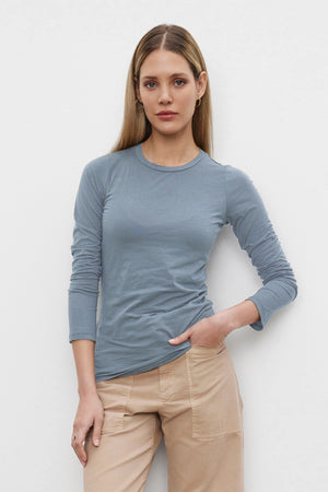 A woman with long blonde hair wearing the ZOFINA TEE from Velvet by Graham & Spencer, a light blue fitted long sleeve tee, along with beige pants stands against a plain white background.