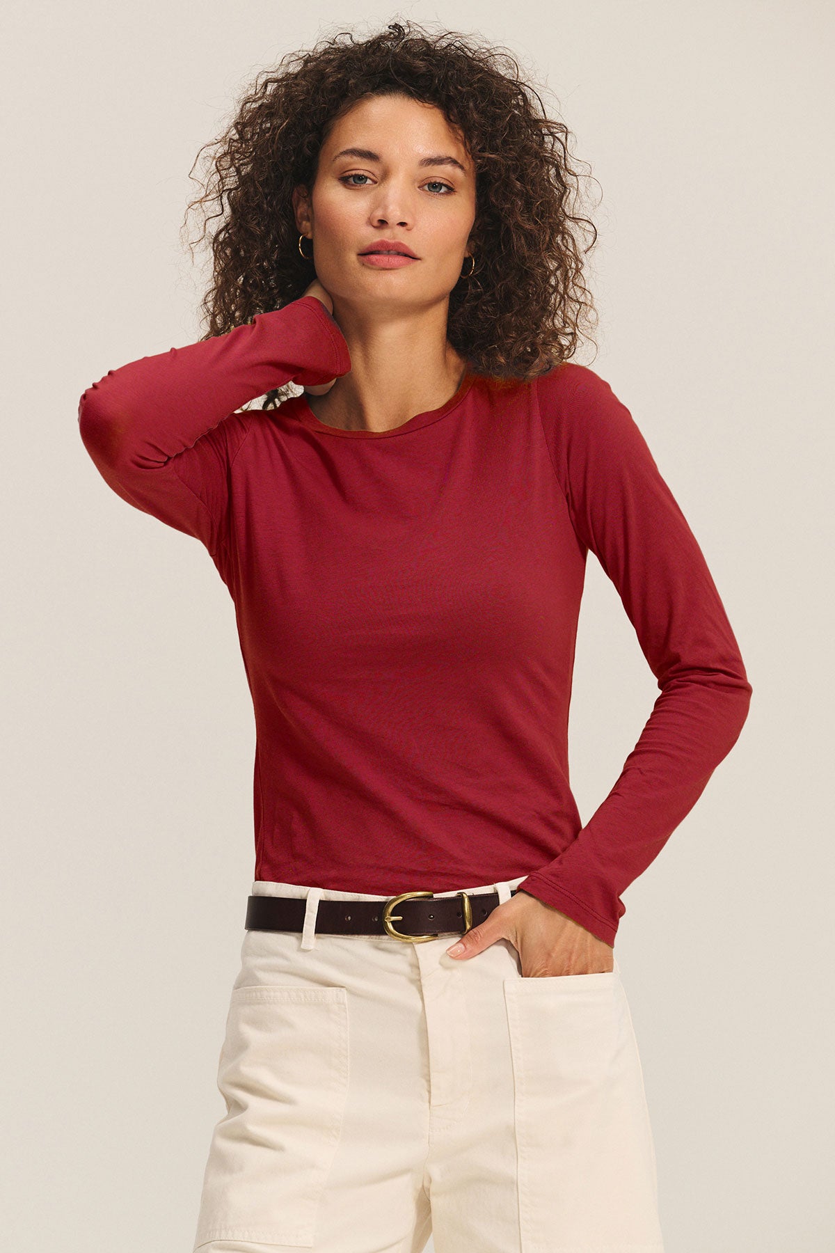 A woman with curly hair wears the ZOFINA TEE by Velvet by Graham & Spencer—an ultra-soft, fitted long sleeve crewneck—tucked into high-waisted white pants with a black belt, standing against a plain background.