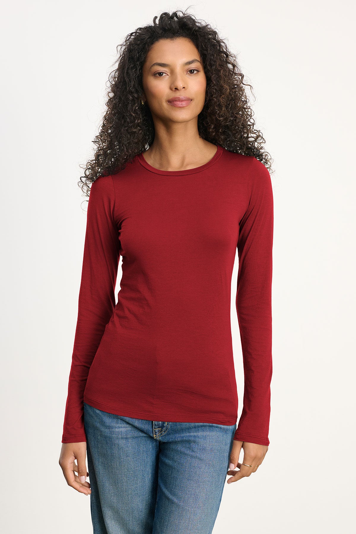 A woman with curly hair wears the ZOFINA TEE by Velvet by Graham & Spencer—a fitted, ultra-soft long sleeve crewneck—paired with blue jeans, standing facing the camera against a plain background.