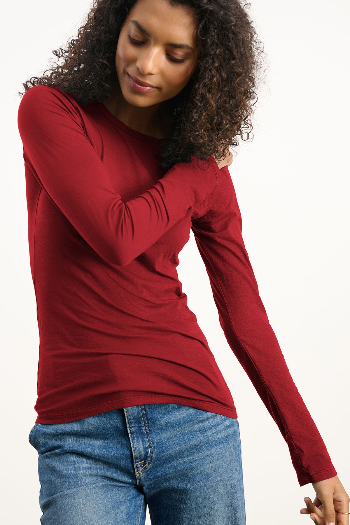 Wearing the ZOFINA TEE by Velvet by Graham & Spencer in ultra-soft red, paired with blue jeans, a person stands against a plain light background, gazing down and bending one arm across their chest.-40417102299329
