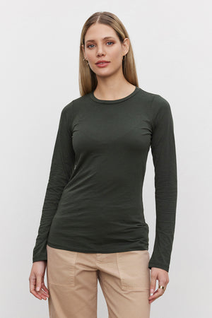 A woman with long blonde hair is wearing a fitted long-sleeve ZOFINA TEE in dark green from Velvet by Graham & Spencer, made with an ultra-soft gauzy whisper fabrication. She pairs it with beige pants and stands against a plain white background.