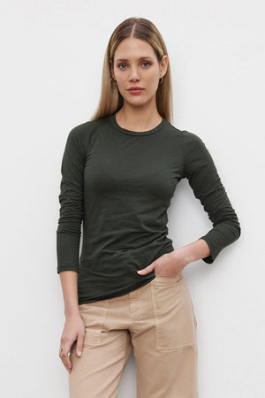 A woman with long, straight hair is standing against a plain white background, wearing the ZOFINA TEE from Velvet by Graham & Spencer in dark green and beige pants, with one hand in her pocket. The fitted long sleeve tee features an ultra-soft gauzy whisper fabric.