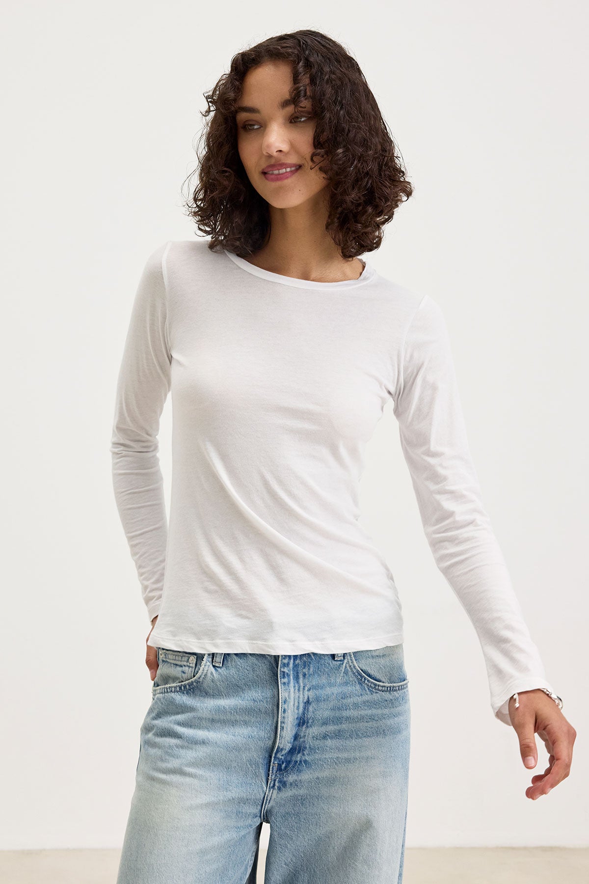   A person with curly hair is wearing the ZOFINA TEE by Velvet by Graham & Spencer, a fitted long-sleeve top with a classic crew neckline, and blue jeans, standing against a plain background. 