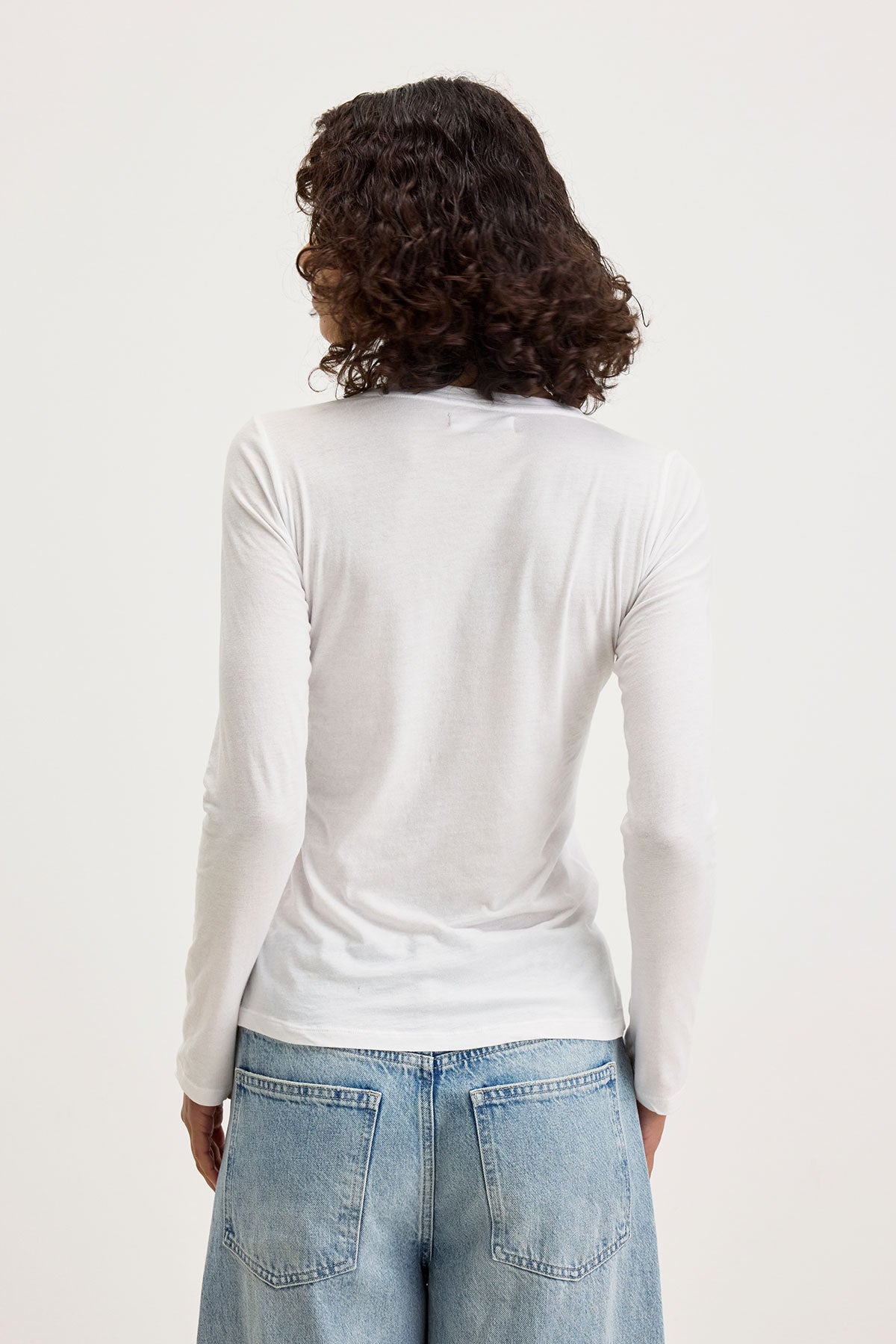   Rear view of a person with curly hair wearing an ultra-soft, fitted long sleeve ZOFINA TEE from Velvet by Graham & Spencer, made from gauzy whisper fabrication, and light blue denim jeans, standing against a plain background. 