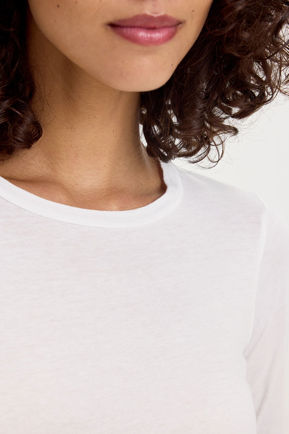   Close-up image of a person wearing the ZOFINA TEE by Velvet by Graham & Spencer. The ultra-soft gauzy whisper fabrication and white crew neck detail are highlighted, with only the lower face, neck, and shoulder area visible. The person's lips are slightly parted, and they have curly hair. 