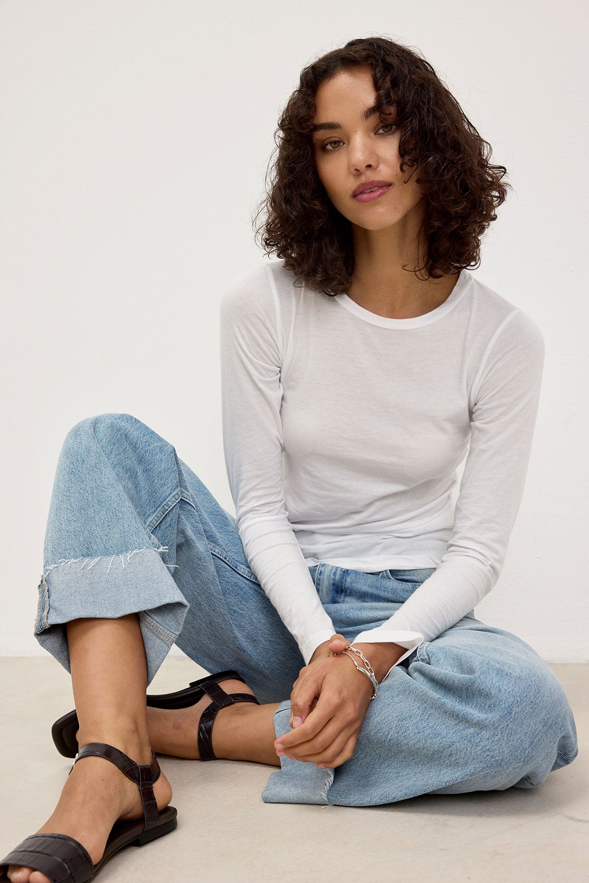   A person with curly hair wearing the ZOFINA TEE from Velvet by Graham & Spencer, paired with blue jeans, sits on the floor with one leg crossed, looking at the camera. 