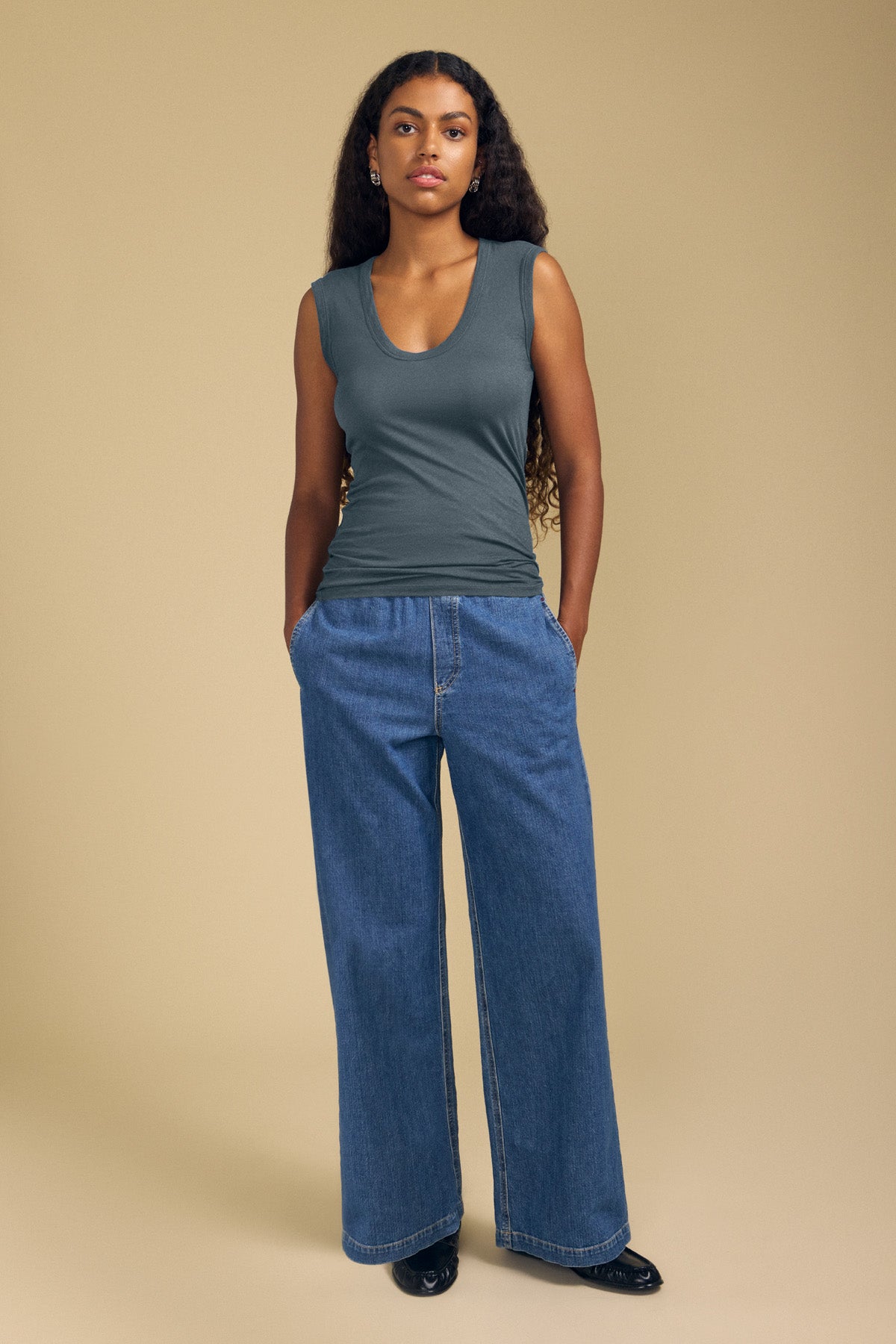 A person stands against a beige background wearing the ESTINA TANK TOP by Velvet by Graham & Spencer—a gauzy cotton tank with a scoop neckline—paired with wide-leg blue jeans and black shoes, hands in pockets for an easy, effortlessly stylish look.-42118984958145