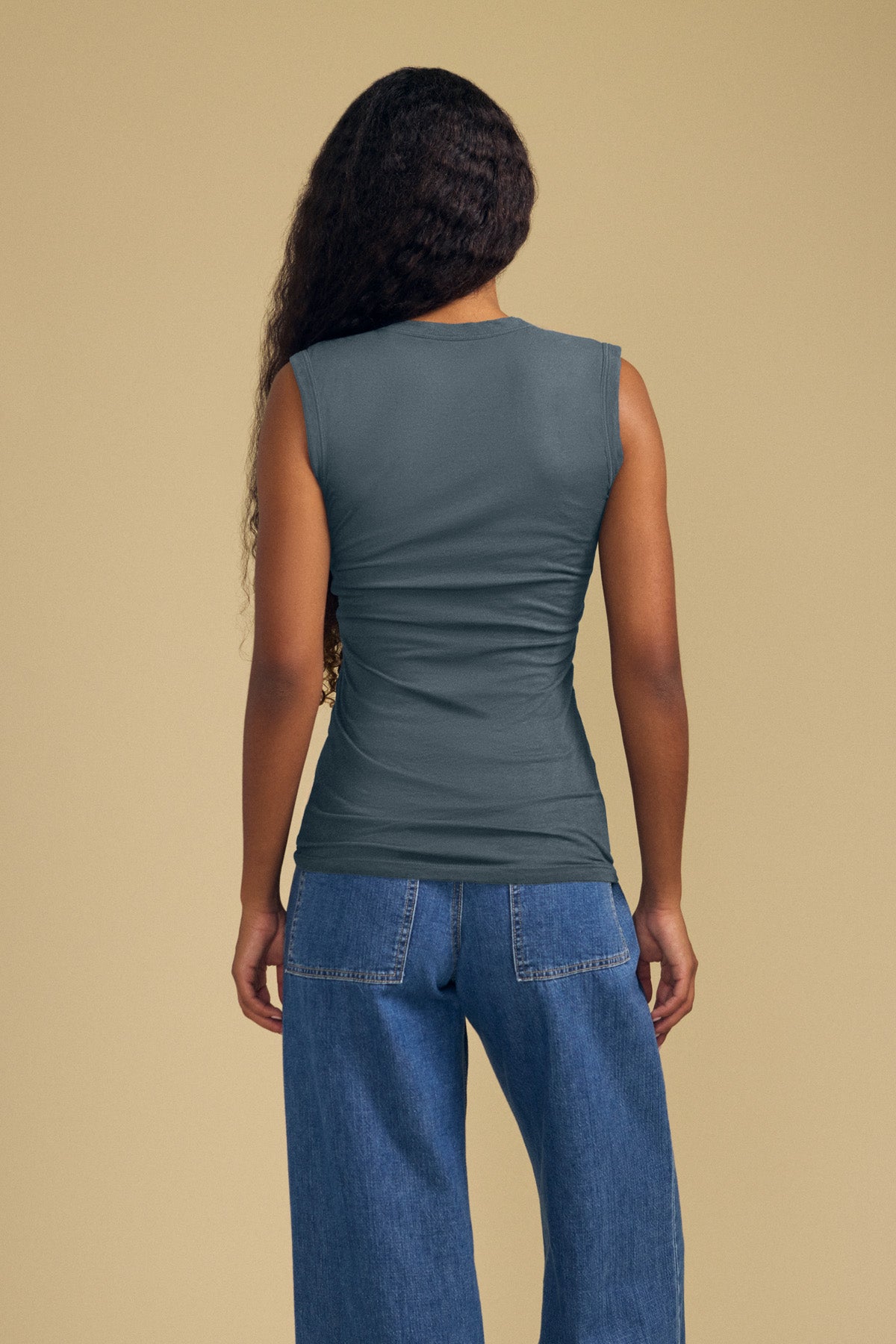 A person stands with their back to the camera, wearing the ESTINA TANK TOP by Velvet by Graham & Spencer—a sleeveless gray gauzy cotton tank with a scoop neckline—paired with blue jeans, set against a plain beige background.