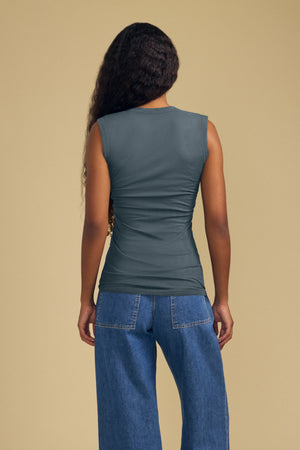A person stands with their back to the camera, wearing the ESTINA TANK TOP by Velvet by Graham & Spencer—a sleeveless gray gauzy cotton tank with a scoop neckline—paired with blue jeans, set against a plain beige background.
