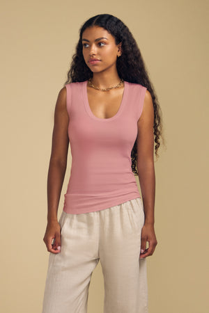 A woman with long curly hair wears the Velvet by Graham & Spencer ESTINA Tank Top—a gauzy cotton pink laid-back tank featuring a flattering scoop neckline—paired with light beige pants, standing against a plain beige background.