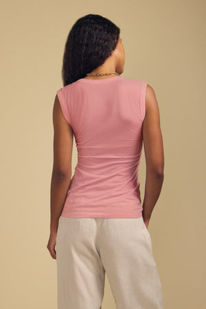 A person stands with their back to the camera, effortlessly styled in the ESTINA TANK TOP by Velvet by Graham & Spencer—a sleeveless, pink gauzy cotton tank featuring a scoop neckline—paired with light-colored pants against a plain beige background.