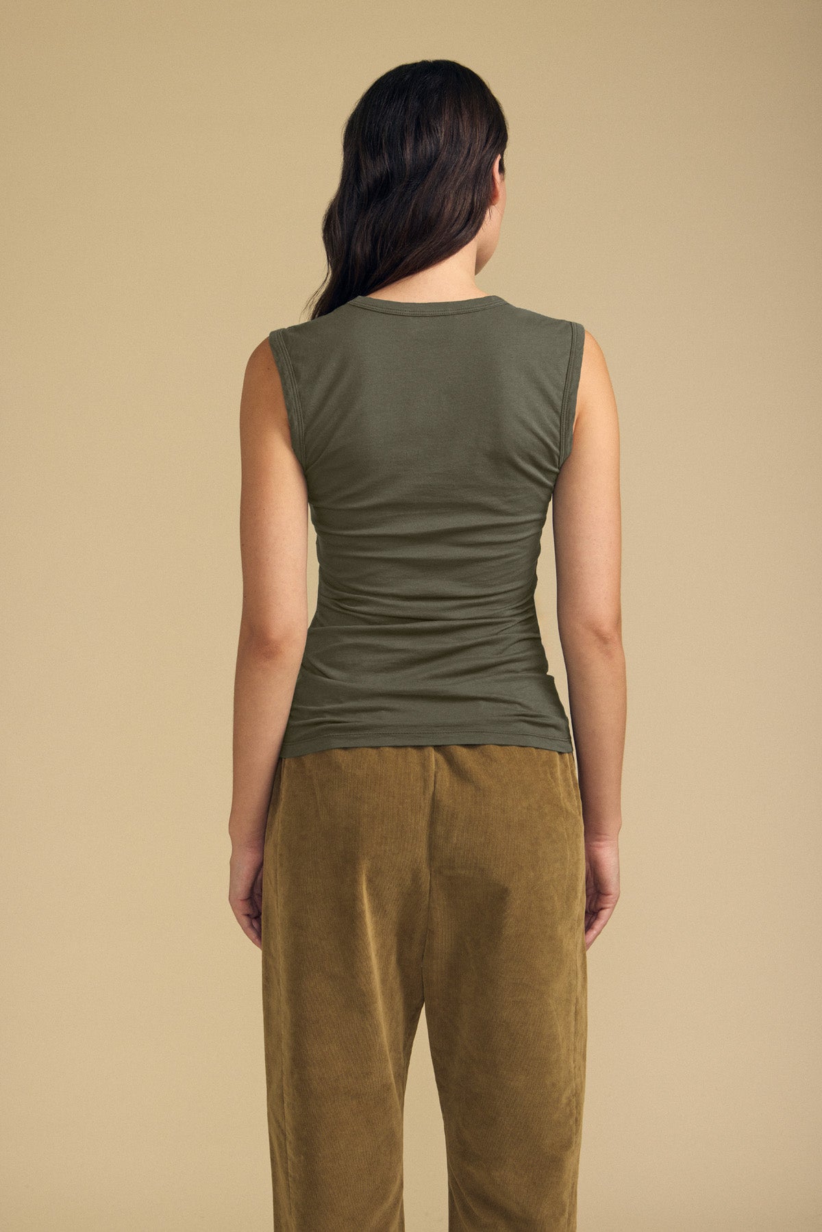 A person stands with their back to the camera, wearing the ESTINA TANK TOP by Velvet by Graham & Spencer in olive green cotton gauze with a scoop neckline, paired with brown pants against a plain beige background.-42118997835969