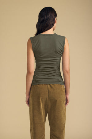 A person stands with their back to the camera, wearing the ESTINA TANK TOP by Velvet by Graham & Spencer in olive green cotton gauze with a scoop neckline, paired with brown pants against a plain beige background.