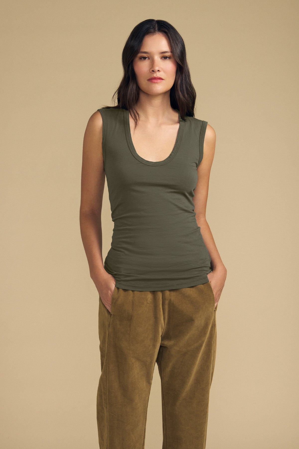 A woman stands against a beige background wearing the ESTINA TANK TOP by Velvet by Graham & Spencer—an olive green gauzy cotton tank featuring a scoop neckline—paired with tan velvet pants, hands in pockets and a neutral expression, embodying effortless style.