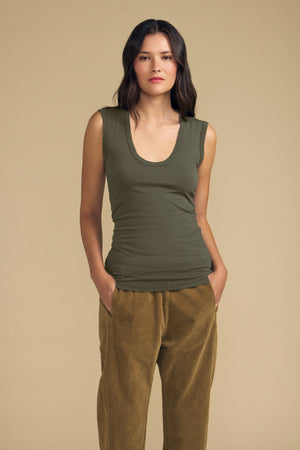 A woman stands against a beige background wearing the ESTINA TANK TOP by Velvet by Graham & Spencer—an olive green gauzy cotton tank featuring a scoop neckline—paired with tan velvet pants, hands in pockets and a neutral expression, embodying effortless style.