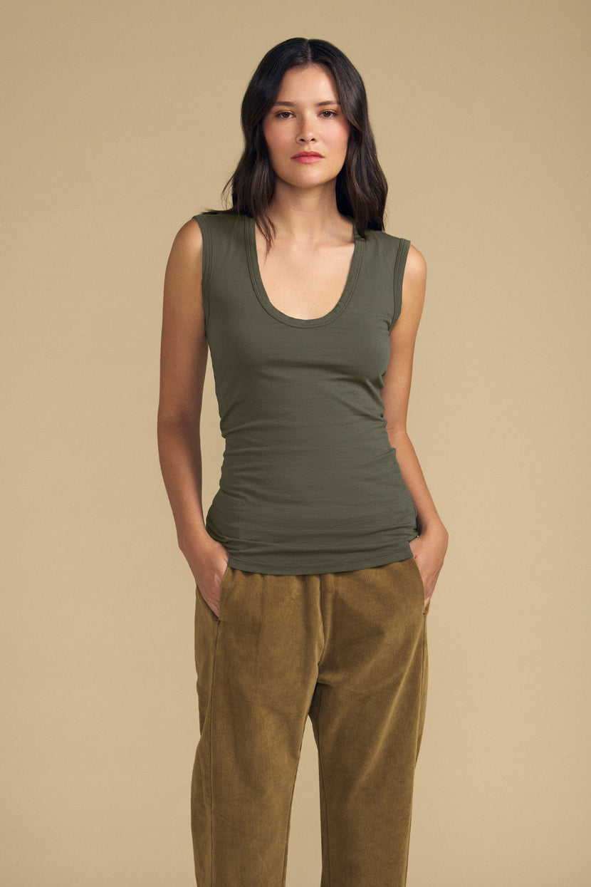 A woman stands against a beige background wearing the ESTINA TANK TOP by Velvet by Graham & Spencer—an olive green gauzy cotton tank featuring a scoop neckline—paired with tan velvet pants, hands in pockets and a neutral expression, embodying effortless style.