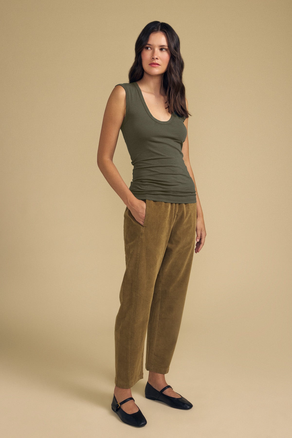 A woman stands against a beige background wearing the ESTINA TANK TOP by Velvet by Graham & Spencer—an olive green, sleeveless top with a scoop neckline—paired with tan pants and black shoes, her hands in her pockets.-42118997737665
