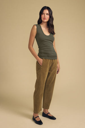 A woman stands against a beige background wearing the ESTINA TANK TOP by Velvet by Graham & Spencer—an olive green, sleeveless top with a scoop neckline—paired with tan pants and black shoes, her hands in her pockets.