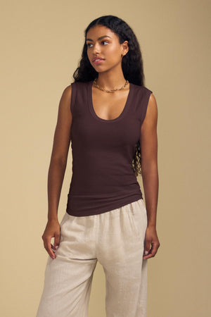 A woman in the ESTINA TANK TOP by Velvet by Graham & Spencer and beige pants stands against a plain beige background, looking to the side.