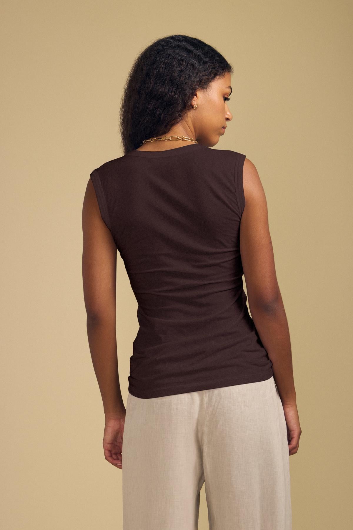 A woman faces away, wearing the ESTINA TANK TOP by Velvet by Graham & Spencer and light beige pants against a plain beige background.-46250769940673