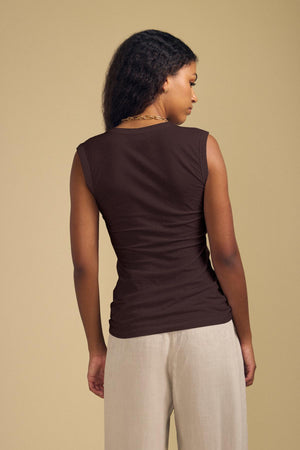 A woman faces away, wearing the ESTINA TANK TOP by Velvet by Graham & Spencer and light beige pants against a plain beige background.