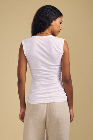 A woman stands facing away from the camera, wearing the ESTINA TANK TOP by Velvet by Graham & Spencer—a white sleeveless top with a scoop neckline—paired with beige pants against a plain beige background.