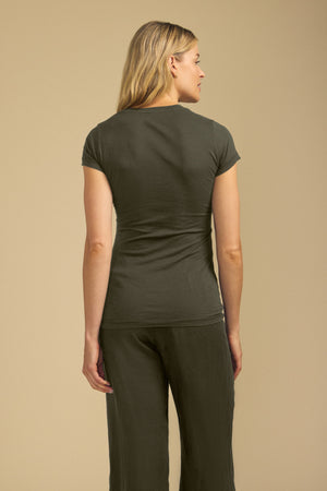A person with light hair stands facing away from the camera, wearing the ultra-soft, short-sleeve JEMMA TEE in olive green by Velvet by Graham & Spencer and matching pants against a beige background.