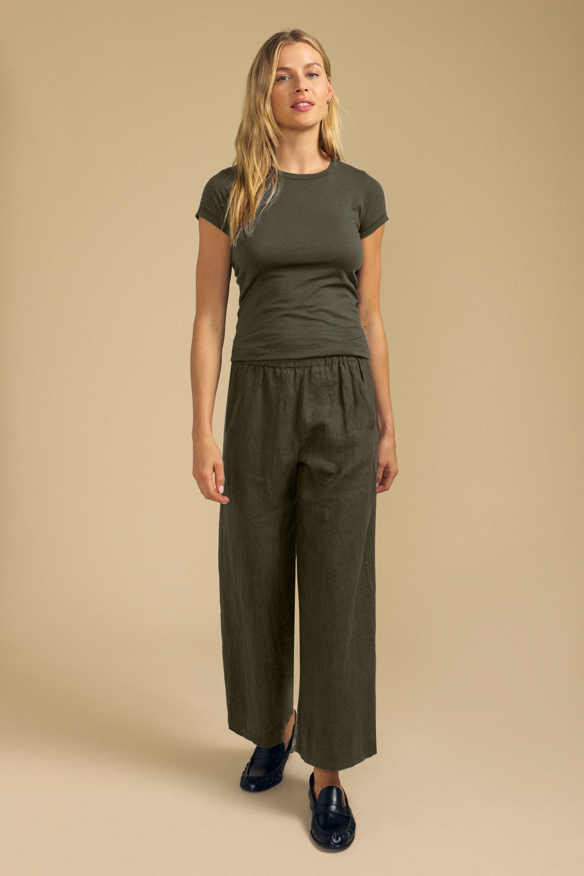 A woman stands against a beige background wearing the JEMMA TEE by Velvet by Graham & Spencer, an olive green fitted top crafted from ultra-soft fabrication, paired with wide-leg pants and black slip-on shoes.-42119196246209