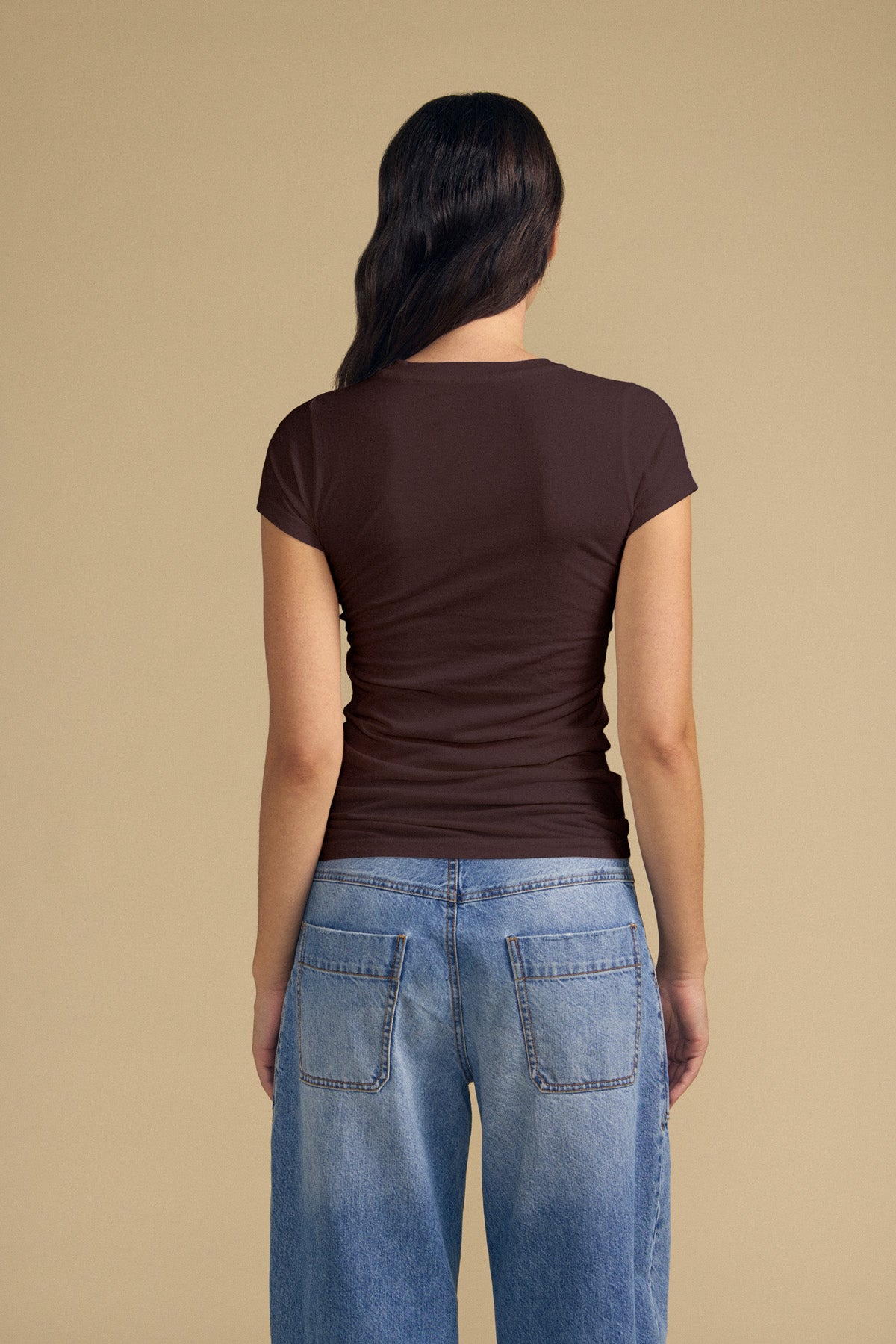 A person with long dark hair, wearing the JEMMA TEE by Velvet by Graham & Spencer in a fitted brown ultra-soft style and loose blue jeans, is standing with their back to the camera against a beige background.-42119021199553