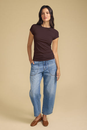 A person stands against a beige backdrop, wearing the Velvet by Graham & Spencer JEMMA TEE—an ultra-soft brown crew neck with short sleeves—paired with loose blue jeans featuring visible stitching and brown flat shoes, with one hand in their pocket.