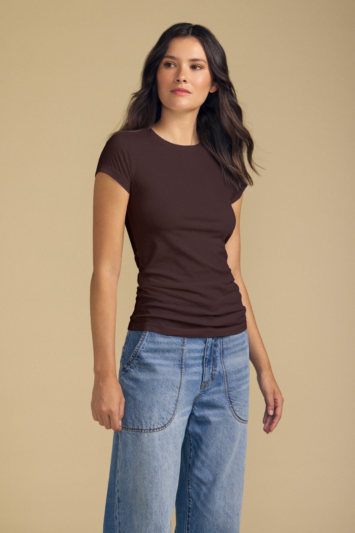 A woman with long brown hair, wearing the Velvet by Graham & Spencer JEMMA TEE—a fitted crew neck top in dark brown—paired with light blue jeans, stands in front of a beige background.-42119021134017