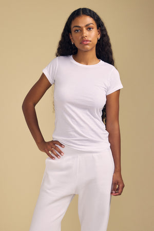 A person with long curly hair wears the JEMMA TEE by Velvet by Graham & Spencer paired with white pants, standing against a beige background with a neutral expression.