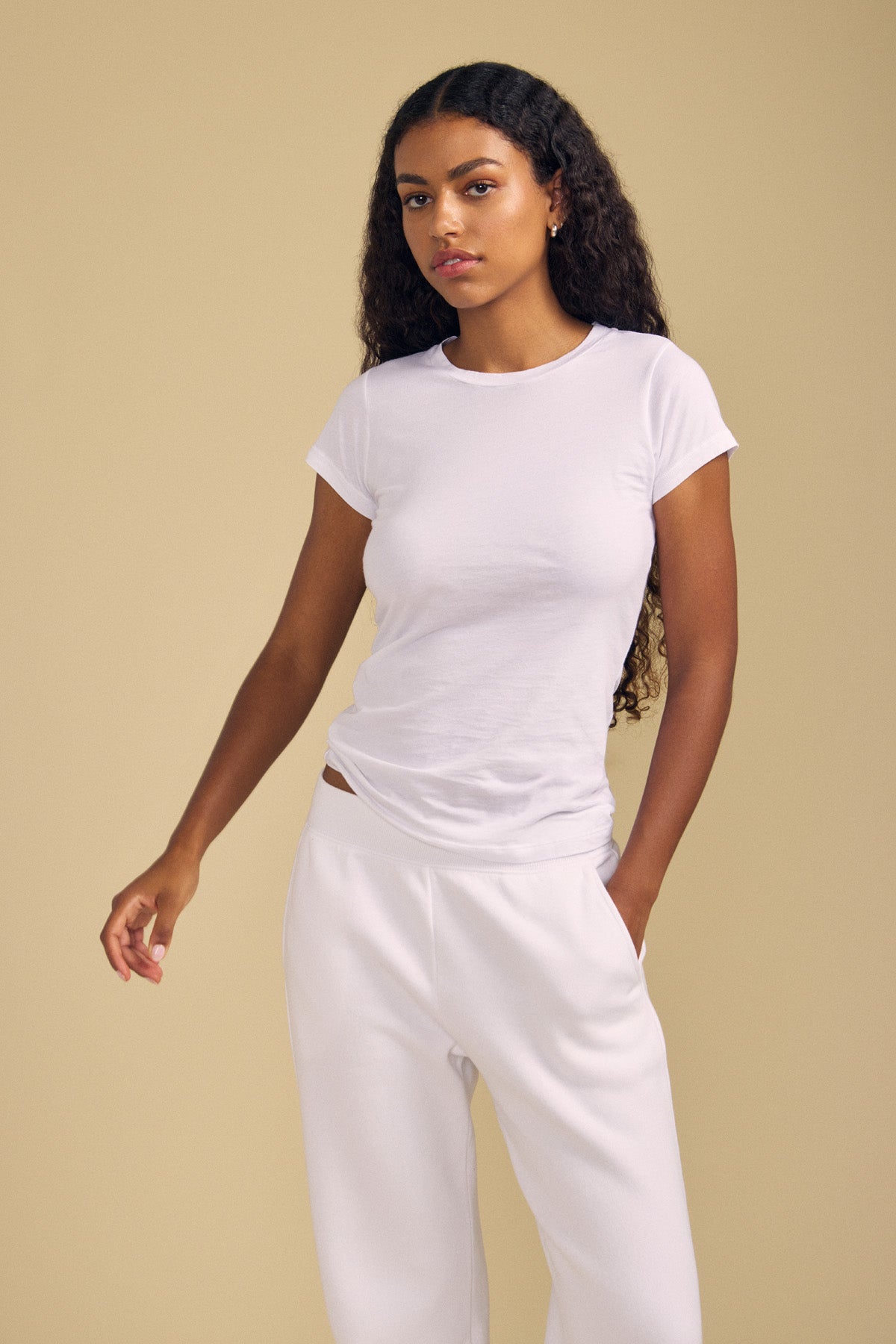 A woman with long curly hair stands against a plain beige background, wearing the fitted white JEMMA TEE by Velvet by Graham & Spencer and white pants, with one hand in her pocket.