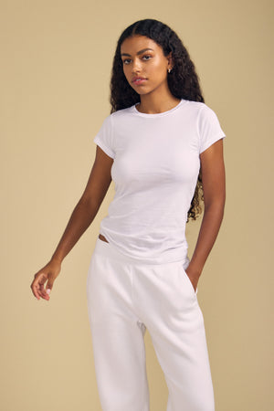 A woman with long curly hair stands against a plain beige background, wearing the fitted white JEMMA TEE by Velvet by Graham & Spencer and white pants, with one hand in her pocket.