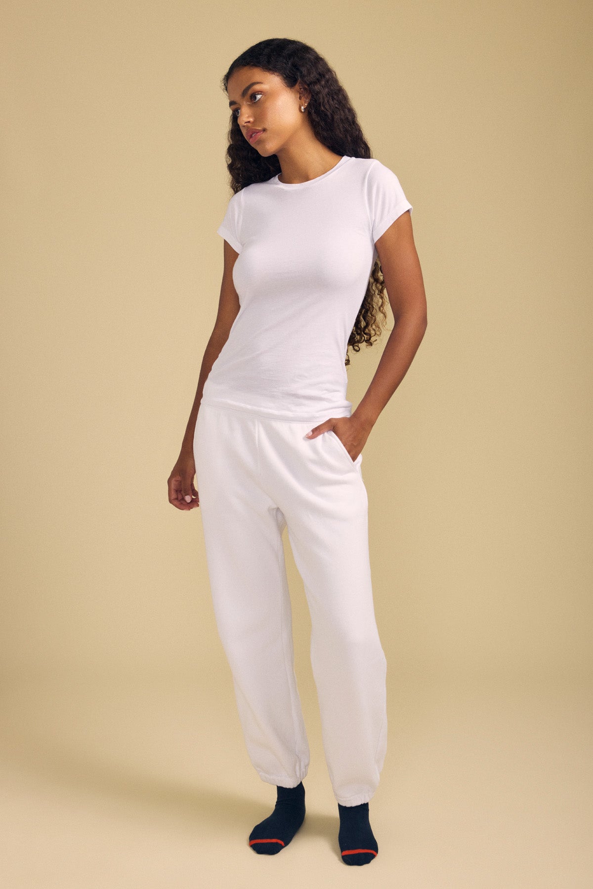 A person stands against a beige background wearing the JEMMA TEE by Velvet by Graham & Spencer, styled with white sweatpants and black socks with red toes.