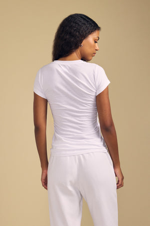 A woman stands facing away from the camera, wearing the ultra-soft JEMMA TEE by Velvet by Graham & Spencer—a short-sleeve white crew neck—paired with white pants against a plain beige background.