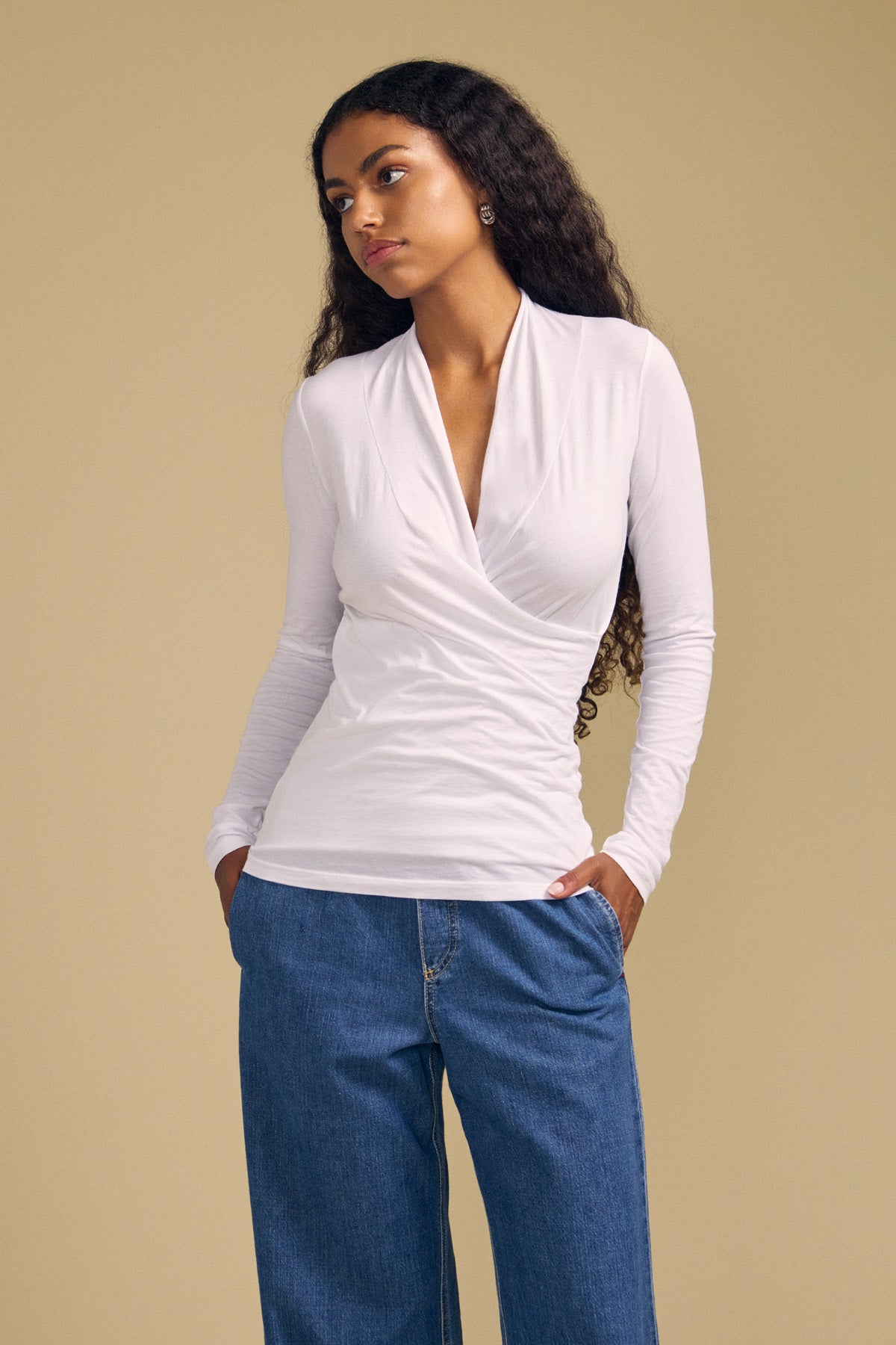 A woman with long curly hair stands with her hands in her pockets against a plain beige background, wearing the MERI TEE by Velvet by Graham & Spencer—a white cross-over wrap tee crafted from lightweight gauzy whisper fabric—paired with blue jeans. The flattering V-neckline of the tee lends an elegant touch to her casual look.-41947059945665