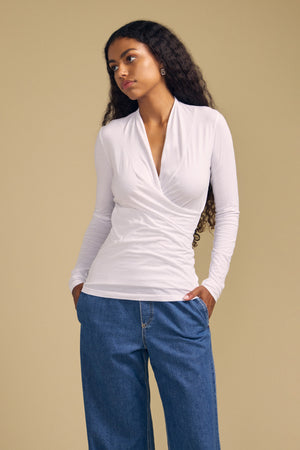 A woman with long curly hair stands with her hands in her pockets against a plain beige background, wearing the MERI TEE by Velvet by Graham & Spencer—a white cross-over wrap tee crafted from lightweight gauzy whisper fabric—paired with blue jeans. The flattering V-neckline of the tee lends an elegant touch to her casual look.
