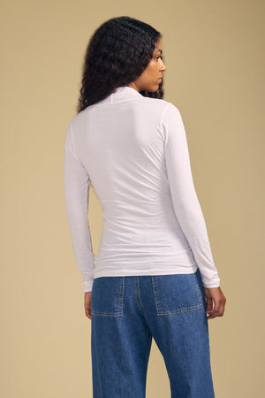 A person stands with their back to the camera, wearing the MERI TEE by Velvet by Graham & Spencer—a cross-over wrap top crafted from lightweight gauzy whisper fabric—paired with blue jeans against a beige background.