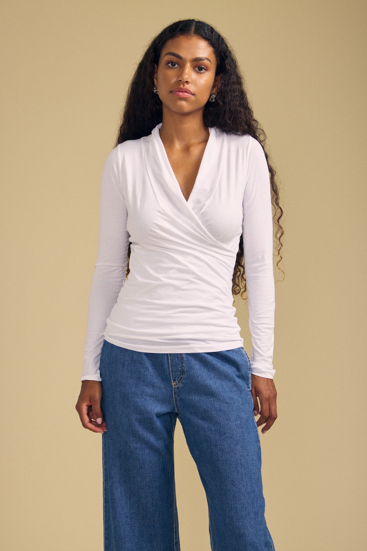 A woman stands against a beige background, wearing the MERI TEE by Velvet by Graham & Spencer—a white long-sleeve cross-over wrap top with a flattering V-neckline—paired with blue jeans. Her long curly hair is worn loose.
